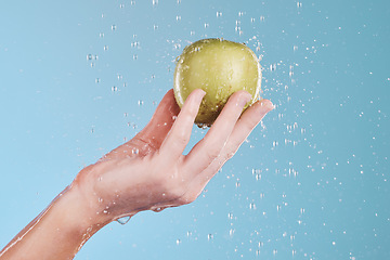 Image showing Apple, cleaning and hand with water drops for wellness and skincare in a studio. Isolated, blue background and nutrition for dermatology, splash and health food with fruit for a diet and healthcare