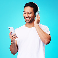 Image showing Headphones, music and Asian man with smartphone for listening in studio isolated on a blue background. Radio, smile and person with mobile, sound or hearing podcast, audio or social media on internet