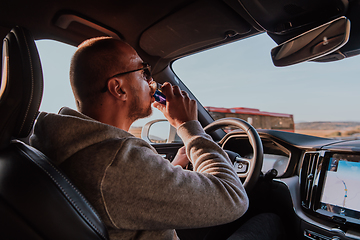 Image showing A tired man drinking acoffee while driving a car at sunset. Tired travel and long drive