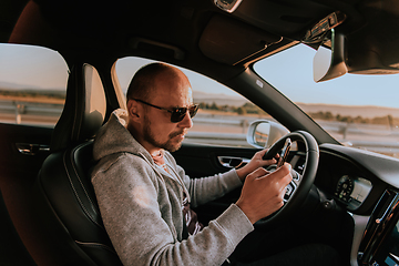 Image showing A man with a sunglasses driving a car and type a message on smartphone at sunset. The concept of car travel