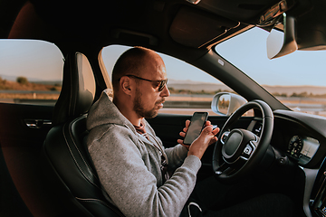 Image showing A man with a sunglasses driving a car and type a message on smartphone at sunset. The concept of car travel