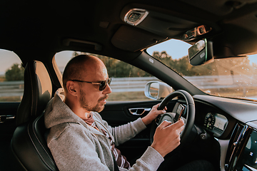Image showing A man with a sunglasses driving a car and type a message on smartphone at sunset. The concept of car travel
