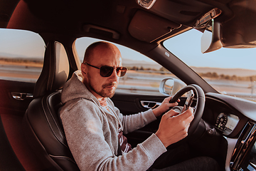 Image showing A man with a sunglasses driving a car and type a message on smartphone at sunset. The concept of car travel