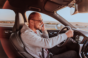 Image showing A man with a sunglasses driving a car and talking on smartphone at sunset. The concept of car travel