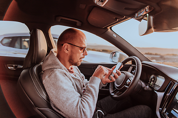 Image showing A man with a sunglasses driving a car and type a message on smartphone at sunset. The concept of car travel