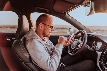 Image showing A man with a sunglasses driving a car and type a message on smartphone at sunset. The concept of car travel