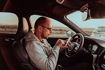 Image showing A man with a sunglasses driving a car and type a message on smartphone at sunset. The concept of car travel