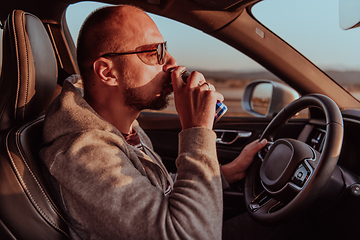 Image showing A tired man drinking acoffee while driving a car at sunset. Tired travel and long drive