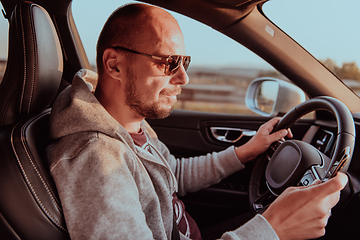 Image showing A man with a sunglasses driving a car and type a message on smartphone at sunset. The concept of car travel