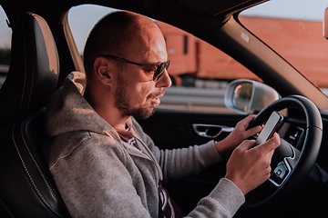 Image showing A man with a sunglasses driving a car and type a message on smartphone at sunset. The concept of car travel