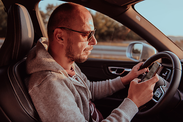 Image showing A man with a sunglasses driving a car and type a message on smartphone at sunset. The concept of car travel