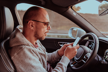 Image showing A man with a sunglasses driving a car and type a message on smartphone at sunset. The concept of car travel