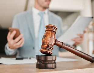 Image showing Closeup, gavel and law firm on table, office or attorney man in blurred background, phone texting or networking. Advocate, lawyer or judge with legal hammer, reading and documents for court evidence