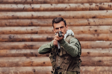 Image showing A soldier in uniform with a rifle in his hand is standing in front of a wooden wall. A soldier guards the forest base from the enemy