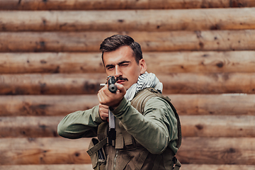 Image showing A soldier in uniform with a rifle in his hand is standing in front of a wooden wall. A soldier guards the forest base from the enemy