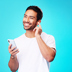 Image showing Asian man, earphones and smartphone in studio for music, podcast app and blue background. Happy male model thinking with mobile phone, listening to audio and streaming sound of radio media with tech