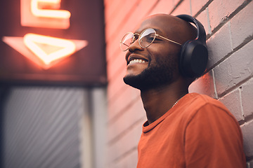 Image showing .Thinking, smile and music with a black man in the city, leaning on a brick wall on the street at night. Idea, glasses and headphones with a happy young male person streaming or listening to audio.