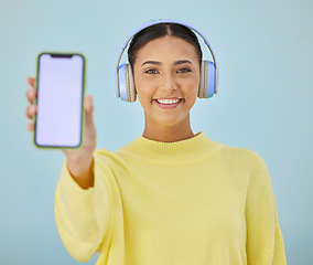 Image showing Portrait of happy woman with phone screen, headphones and mockup in studio for social media, mobile app and streaming radio. Smile, music subscription and girl with cellphone on blue background space