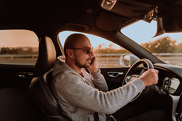 Image showing A man with a sunglasses driving a car and talking on smartphone at sunset. The concept of car travel