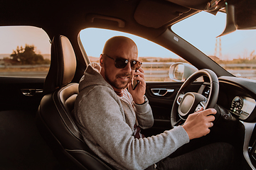 Image showing A man with a sunglasses driving a car and talking on smartphone at sunset. The concept of car travel