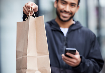 Image showing Courier man, giving paper bag and portrait with phone, smile and delivery service in supply chain job. Young logistics worker, package and hand with smartphone, product and e commerce in metro cbd