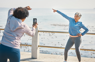 Image showing Phone, friends and photo of senior woman pointing at beach with silly pose at sea for fitness. Exercise, mobile and picture for social media post on a ocean promenade walk for workout and friendship