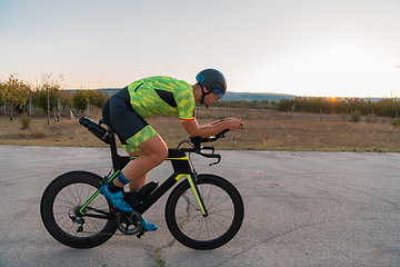 Image showing Triathlete riding his bicycle during sunset, preparing for a marathon. The warm colors of the sky provide a beautiful backdrop for his determined and focused effort.