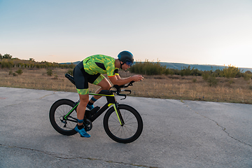 Image showing Triathlete riding his bicycle during sunset, preparing for a marathon. The warm colors of the sky provide a beautiful backdrop for his determined and focused effort.