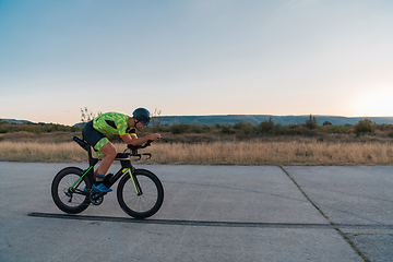 Image showing Triathlete riding his bicycle during sunset, preparing for a marathon. The warm colors of the sky provide a beautiful backdrop for his determined and focused effort.