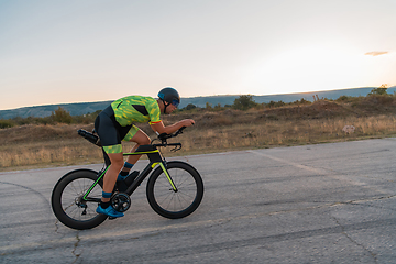 Image showing Triathlete riding his bicycle during sunset, preparing for a marathon. The warm colors of the sky provide a beautiful backdrop for his determined and focused effort.