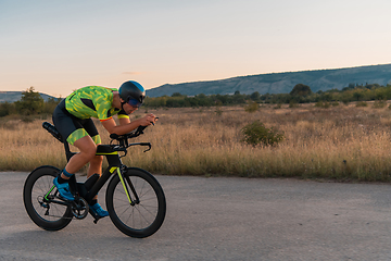 Image showing Triathlete riding his bicycle during sunset, preparing for a marathon. The warm colors of the sky provide a beautiful backdrop for his determined and focused effort.