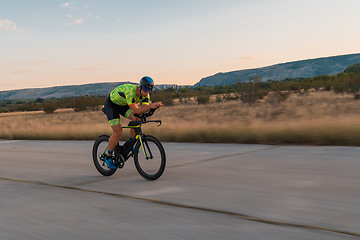 Image showing Triathlete riding his bicycle during sunset, preparing for a marathon. The warm colors of the sky provide a beautiful backdrop for his determined and focused effort.