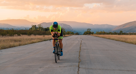 Image showing Triathlete riding his bicycle during sunset, preparing for a marathon. The warm colors of the sky provide a beautiful backdrop for his determined and focused effort.