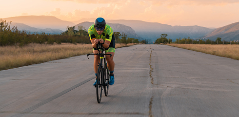 Image showing Triathlete riding his bicycle during sunset, preparing for a marathon. The warm colors of the sky provide a beautiful backdrop for his determined and focused effort.