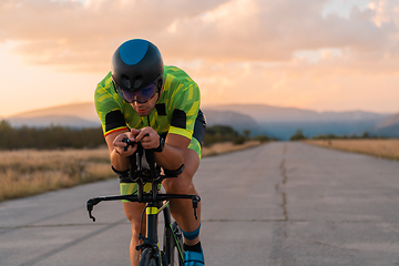 Image showing Triathlete riding his bicycle during sunset, preparing for a marathon. The warm colors of the sky provide a beautiful backdrop for his determined and focused effort.