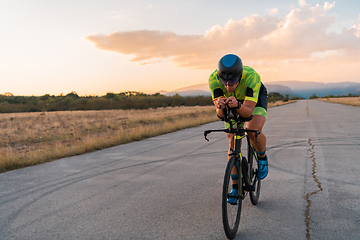 Image showing Triathlete riding his bicycle during sunset, preparing for a marathon. The warm colors of the sky provide a beautiful backdrop for his determined and focused effort.