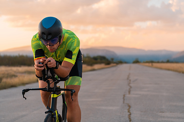 Image showing Triathlete riding his bicycle during sunset, preparing for a marathon. The warm colors of the sky provide a beautiful backdrop for his determined and focused effort.