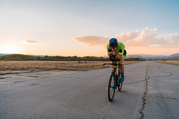 Image showing Triathlete riding his bicycle during sunset, preparing for a marathon. The warm colors of the sky provide a beautiful backdrop for his determined and focused effort.