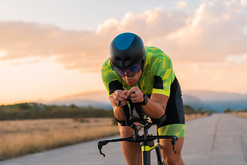 Image showing Triathlete riding his bicycle during sunset, preparing for a marathon. The warm colors of the sky provide a beautiful backdrop for his determined and focused effort.