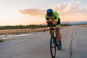 Image showing Triathlete riding his bicycle during sunset, preparing for a marathon. The warm colors of the sky provide a beautiful backdrop for his determined and focused effort.