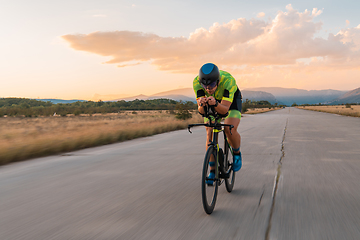 Image showing Triathlete riding his bicycle during sunset, preparing for a marathon. The warm colors of the sky provide a beautiful backdrop for his determined and focused effort.