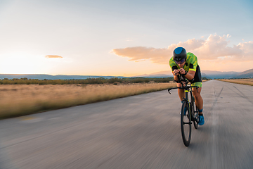 Image showing Triathlete riding his bicycle during sunset, preparing for a marathon. The warm colors of the sky provide a beautiful backdrop for his determined and focused effort.