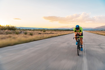 Image showing Triathlete riding his bicycle during sunset, preparing for a marathon. The warm colors of the sky provide a beautiful backdrop for his determined and focused effort.