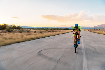 Image showing Triathlete riding his bicycle during sunset, preparing for a marathon. The warm colors of the sky provide a beautiful backdrop for his determined and focused effort.