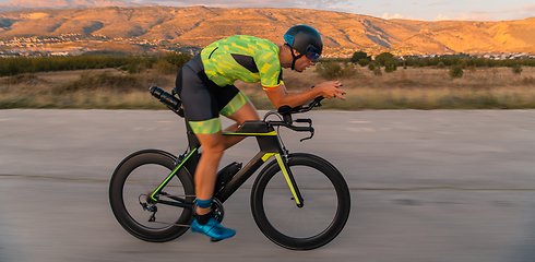 Image showing Triathlete riding his bicycle during sunset, preparing for a marathon. The warm colors of the sky provide a beautiful backdrop for his determined and focused effort.