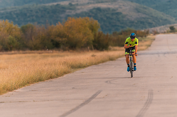 Image showing Triathlete riding his bicycle during sunset, preparing for a marathon. The warm colors of the sky provide a beautiful backdrop for his determined and focused effort.