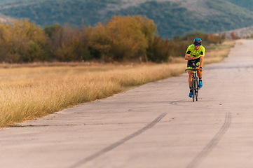 Image showing Triathlete riding his bicycle during sunset, preparing for a marathon. The warm colors of the sky provide a beautiful backdrop for his determined and focused effort.