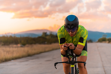 Image showing Close up photo of triathlete riding his bicycle during sunset, preparing for a marathon. The warm colors of the sky provide a beautiful backdrop for his determined and focused effort.