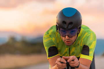 Image showing Close up photo of triathlete riding his bicycle during sunset, preparing for a marathon. The warm colors of the sky provide a beautiful backdrop for his determined and focused effort.