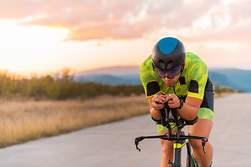 Image showing Close up photo of triathlete riding his bicycle during sunset, preparing for a marathon. The warm colors of the sky provide a beautiful backdrop for his determined and focused effort.